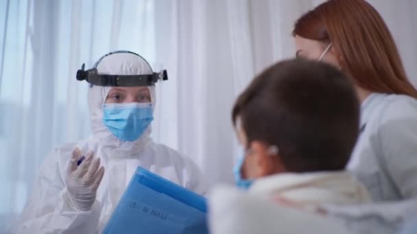 Γυναίκα γιατρός σε προστατευτική στολή με κράνος και ιατρική μάσκα για την προστασία από θανατηφόρα μόλυνση μετρά τη θερμοκρασία με ένα ανέπαφο θερμόμετρο για λίγο αρσενικό ασθενή δίπλα στη μητέρα — Αρχείο Βίντεο