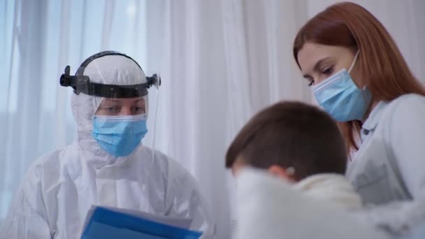 Virüs ve enfeksiyondan korunmak için güvenlik kaskı, tıbbi maske takan doktor bir hastayı muayene ederken genç bir çocuğa beş tane veriyor. — Stok video