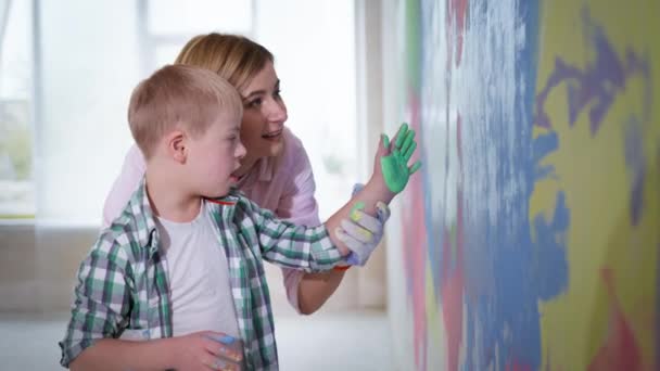 Ocio familiar juntos en el nuevo hogar, niño con síndrome de Down con pintura en la palma de la mano pone la impresión en una pared de color con la ayuda de mamá — Vídeo de stock