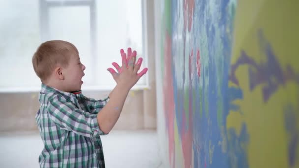 Дитячі розваги, хлопчик з синдромом вниз і дівчина з фарбою на долонях кладе руки на кольорову стіну під час ремонту в новій квартирі — стокове відео