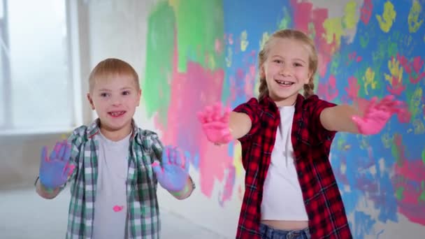 Crianças brincando com cores em casa durante a renovação, menina com aparelho e menino com síndrome de down sorrindo mostrando à câmera coloridas palmas pintadas — Vídeo de Stock