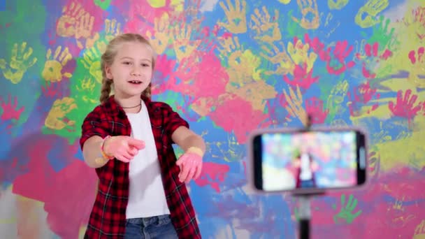 Детское развлечение, портрет девочки в брекетах во время ремонта с цветными руками в красках снимает видеоблог на камеру мобильного телефона — стоковое видео