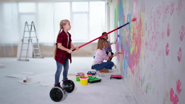 Criança em transporte elétrico com rolo rosa pinta as paredes na sala, menina ajuda os pais durante a renovação — Vídeo de Stock