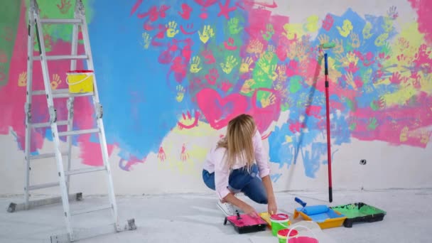 Artista chica sumerge sus manos en bandeja con pintura durante una renovación del hogar inusual, mujer joven crea un cuadro en la pared cerca de escalera interior — Vídeo de stock