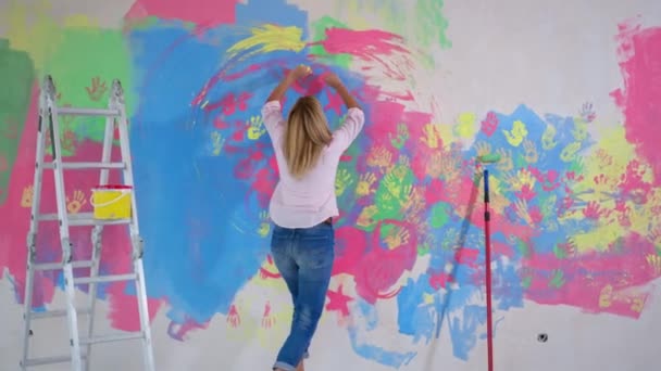 Ζωγράφος δημιουργεί σύγχρονη τέχνη, κορίτσι βουτάει τα χέρια της σε δίσκο με μπογιά στη συνέχεια ζωγραφίζει με παλάμες στον τοίχο κατά τη διάρκεια της ανακαίνισης, πίσω όψη — Αρχείο Βίντεο