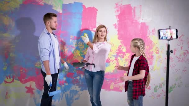 Blog during renovation, girl with parents paint the walls with roller and brush at camera of mobile phone — Vídeo de Stock
