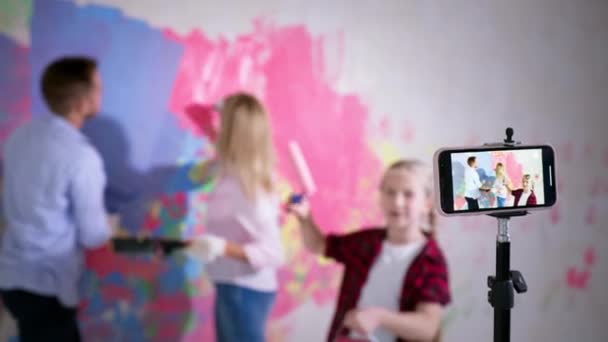 Vloggning och renovering, barn flicka med tandställning talar i kameran på bakgrund av föräldrar med bricka och penslar måla väggen i olika färger — Stockvideo