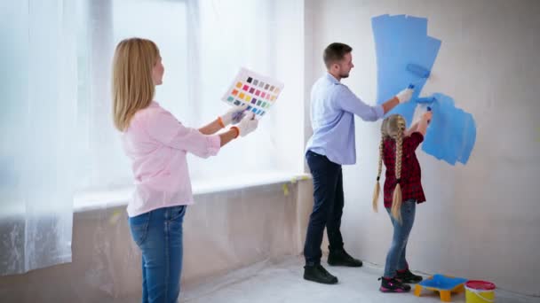 Ház szoba felújítás, fiatal család mutatja, hogyan kell festeni falak görgők, amelyek borított kék festék az imádnivaló lánya — Stock videók