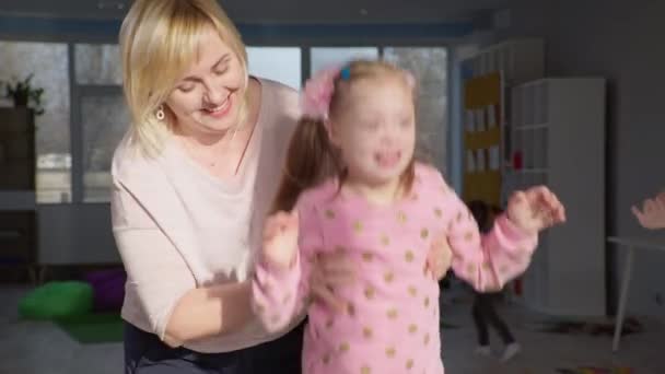 Маленька дівчинка з синдромом Дауна стрибає на батуті і плескає руками, жінка-вчителька підтримує щасливу дитину з інвалідністю під час активних ігор — стокове відео