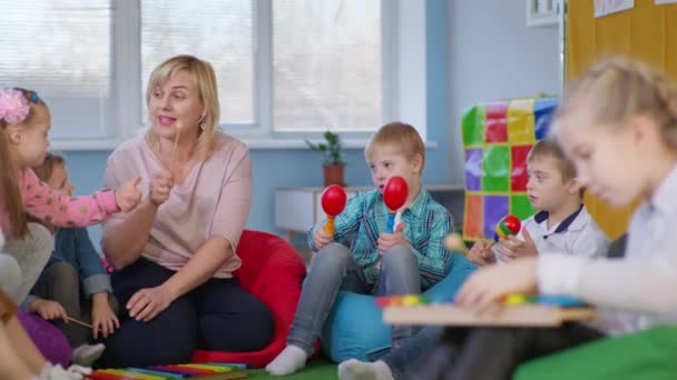 Bambini tempo libero, i ragazzi con disabilità giocano sulle maracas con i bambini e l'insegnante durante la lezione di musica seduti su sedie multicolore sacchetto di fagioli — Video Stock