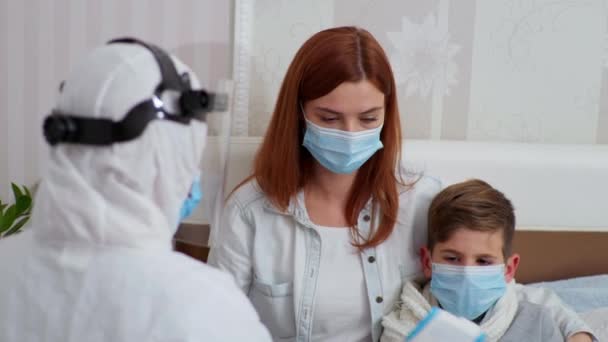 Pracovník v ochranném obleku proti viru a infekci poskytuje lékařskou péči matce s dětskou maskou na obličeji a měří tělesnou teplotu bezkontaktním teploměrem ve stupních Faryngate — Stock video