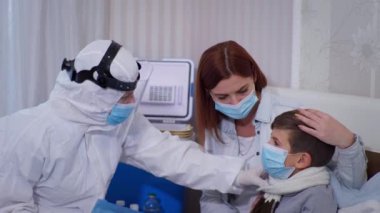 Tehlikeli virüse karşı korunmak için koruyucu giysi ve maske takan sağlık çalışanı koronavirüs semptomlarına sahip erkek çocuğu muayene ediyor.