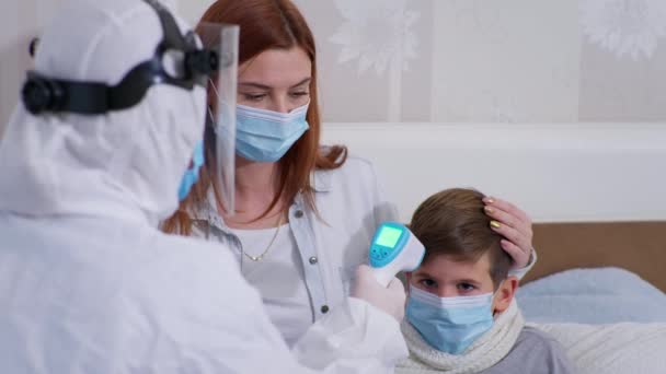 Médico femenino en un traje de protección examina la salud de un niño pequeño con síntomas de un virus y la infección y muestra a su madre en una máscara facial en un termómetro sin contacto — Vídeos de Stock