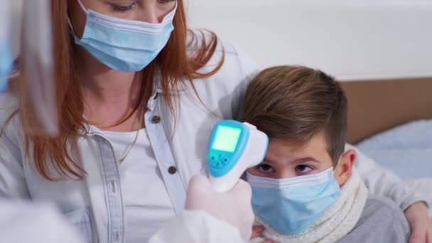 Ärztin im Schutzanzug untersucht kleine Patientin zu Hause und misst Körpertemperatur mit einem berührungslosen Thermometer in Celsius — Stockvideo
