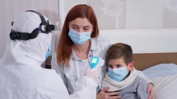 Virüs ve enfeksiyona karşı korunmak için koruyucu giysi ve maske takan hemşire temas olmayan termometreye sahip sağlıksız bir erkek çocuğu ölçer ve oğlunun vücut sıcaklığını annesine gösterir. — Stok video