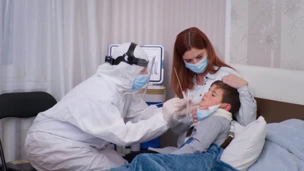 Oficial médico en traje protector examina paciente niño varón y toma la prueba del coronavirus, madre cariñosa cuida de la salud del hijo malsano — Vídeos de Stock