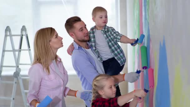 Mujer y hombre con síndrome de Down niño en brazos y mujer niño pintar la pared con pinturas de colores en el interior durante la renovación — Vídeos de Stock