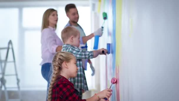 Bella ragazza con rullo rosa nelle mani dipinge pareti sfondo dei genitori e fratello con sindrome di Down durante il lavoro di finitura in casa — Video Stock