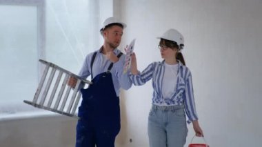 Erkek ve kadın tamirciler basamaklı güvenlik kaskı takıyor ve dallarla boya yapıyorlar. Kapalı alandaki duvarları boyuyorlar.
