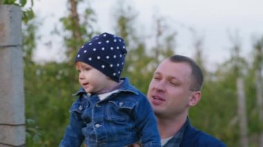 Baba, baba küçük çocuğunu bir meyve bahçesinde aile eğlencesi sırasında yeşil bir ağaca sarılır.