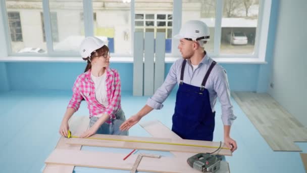 Женщина и мужчина встроенные жесткие шляпы измеряют ламинат с помощью рулетки для ремонта пола в новой квартире — стоковое видео
