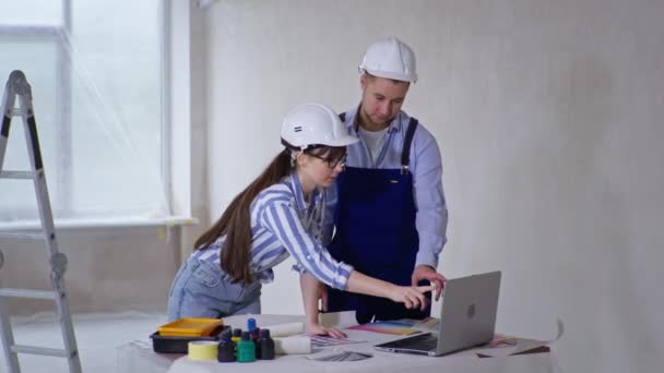 Креативна молода жінка і чоловік в робочому одязі і шоломі обговорюють дизайн кімнати на ноутбуці вибирайте новий тон для стін в квартирі — стокове відео