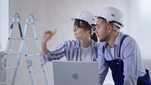 Perempuan dan laki-laki dalam helm sedang mendiskusikan proyek renovasi ruang pada laptop dan memilih warna dinding dengan palet warna di tangan mereka — Stok Video