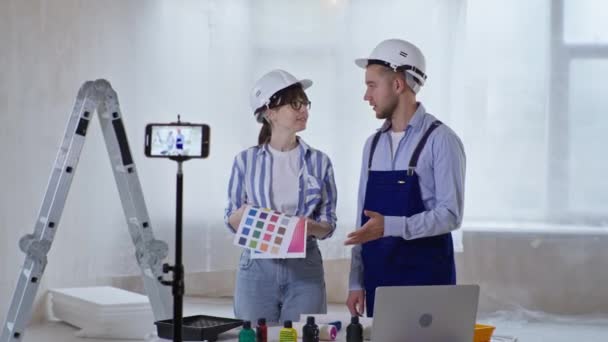 Profesionales masculinos y femeninos hablan sobre la nueva pintura de pared y muestran esquema de color y rodillo de pintura en la cámara del teléfono mientras graban video para el blog en el teléfono inteligente — Vídeos de Stock