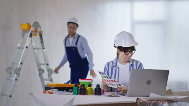 Joven especialista en construcción masculina consulta con el diseñador femenino sentado en el proyecto de renovación en el ordenador portátil sobre el color de las paredes mientras que la pintura — Vídeos de Stock