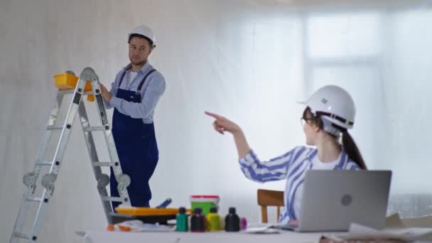 Pretty girl designer sits at laptop with color scheme in her hands and gives instructions to male construction worker — Stock Video