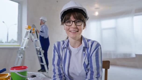 Jeune femme constructeur de casque communique avec la famille via lien vidéo tout en travaillant sur le fond du chantier de construction de mur de peinture homme — Video