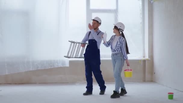 And woman in protective helmets with stepladder and bucket of paint in their hands discussing color of walls — Stock Video