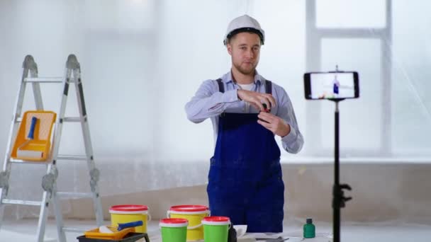 Uomo in casco parla di costruzione di vernici per pareti e punti alla fotocamera su smartphone durante la trasmissione in diretta indoor — Video Stock