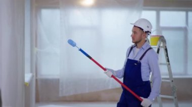 İş elbisesi içindeki erkek boyacı ve güvenlik kaskı bina içinde tamirat yapıyor ve inşaat tekerleği kullanarak duvarları boyuyor.