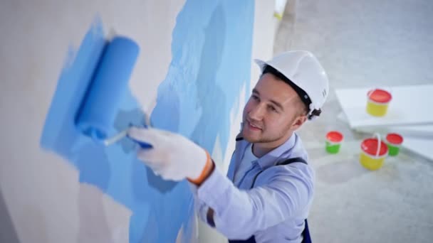 Молодой художник выполняет отделочные работы на строительной площадке и использует ролики для покраски стен — стоковое видео
