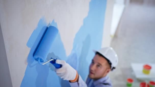 Hombre en sombrero duro realiza el trabajo de acabado y pinta las paredes de color azul con rodillo, durante el reacondicionamiento en interiores — Vídeos de Stock