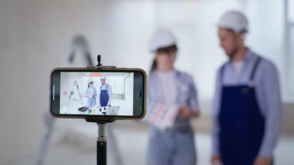 Man and woman doing repairs indoors and recording video on smartphone about painting walls at construction site — Stock Video