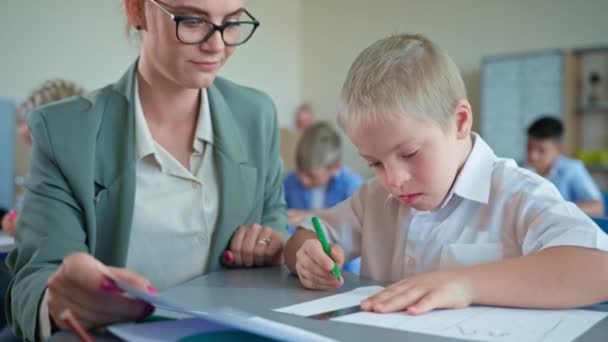 Lehrerin arbeitet mit besonderem Jungen mit Down-Syndrom-Nahaufnahme, junge Frau mit Brille unterrichtet Schülerin am Schreibtisch der Grundschule — Stockvideo