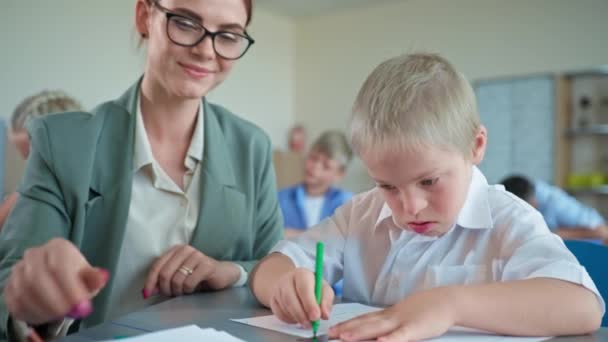 Pedagogo trabaja con síndrome de Down chico, mujer joven en gafas enseña el alumno sentado en el escritorio en el primer plano de la escuela primaria — Vídeo de stock