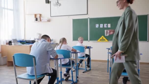 Insegnante premuroso porta un bicchiere d'acqua a un ragazzo a scuola, gli alunni si siedono alla scrivania durante una lezione o pausa in classe — Video Stock