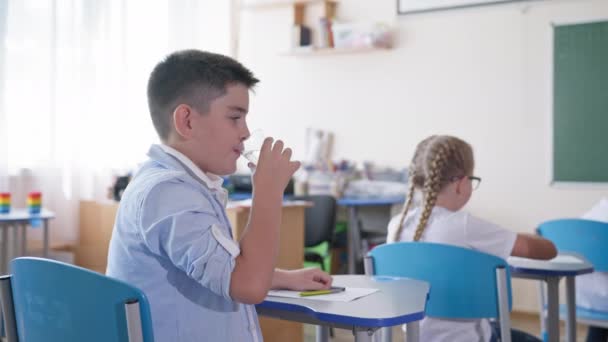 Scolaro bere acqua mentre seduto alla scrivania in classe durante una lezione sullo sfondo dei compagni di classe, scuola elementare — Video Stock