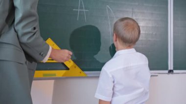 Down sendromlu öğrenci tahtada öğretmenle duruyor ve ilkokulda beyaz tebeşirle yazmayı öğreniyor.