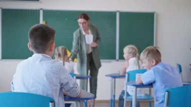 Sınıf arkadaşları ders sırasında birbirleriyle konuşuyorlar, öğretmen öğrenci kartlarını karatahtanın yanına gösteriyor ve okuldaki çocuğa dikiz aynasından soruyor.