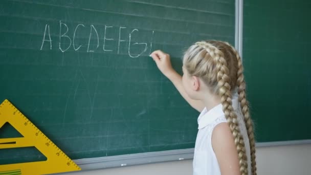 Portrait d'écolière heureuse au tableau noir écrit des lettres avec de la craie blanche en classe et regarde la caméra, leçon d'anglais à l'école primaire — Video