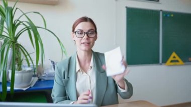 Kameraya bakan gülümseyen öğretmenin portresi, elinde kartlar olan gözlüklü pedagog video dersi kara tahtanın arkasındaki masada oturuyor.