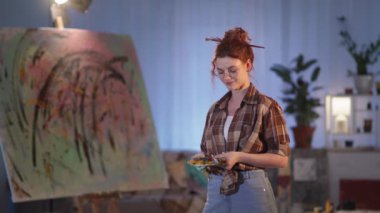 Kadın ressam sehpanın yanında fırça ve palet tutuyor. Yeni boyanmış bir resim. Stüdyoda kameraya gülümseyen mutlu bir kız.