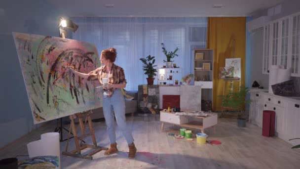 Καλλιτέχνης κορίτσι κάνει τις τελευταίες πινελιές σε καμβά, δημιουργική νεαρή γυναίκα με πινέλα και χρώματα στα χέρια κοιτάζει τη δημιουργία του στο καβαλέτο κατά τη διάρκεια πίνοντας τσάι από ένα φλιτζάνι στο σύγχρονο Wor — Αρχείο Βίντεο