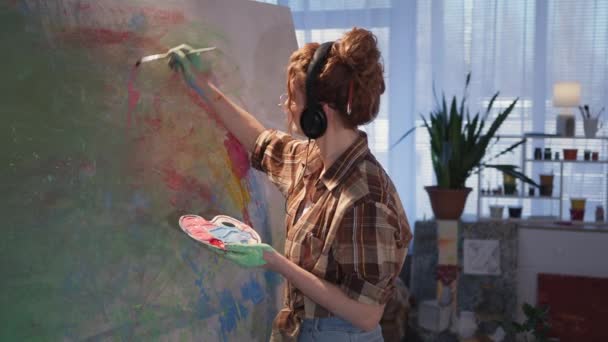 Νεαρή κομψή γυναίκα καλλιτέχνης σε ακουστικά με βρώμικα χέρια στη ζωγραφική εικόνα σε μεγάλο καμβά, κορίτσι με βούρτσα δημιουργώντας έργο τέχνης στο καβαλέτο στο στούντιο — Αρχείο Βίντεο