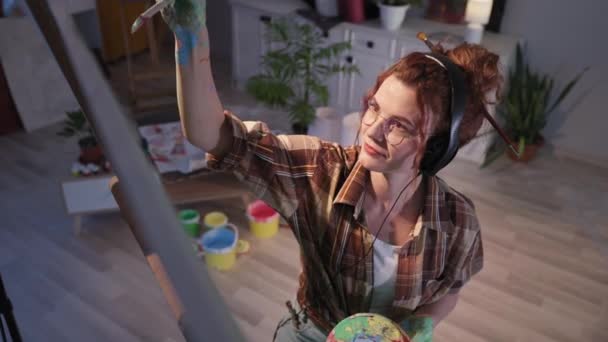 Stylová mladá žena v brýlích se špinavýma rukama vytváří obraz pomocí barev a štětcem, umělec poslouchá hudbu na sluchátkách ve studiu — Stock video