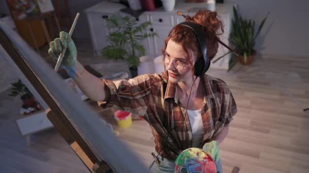 Umělkyně mladá žena poslouchá hudbu na sluchátka a maluje obraz na velké plátno se štětcem, krásná dívka se špinavýma rukama v barvě v dílně — Stock video